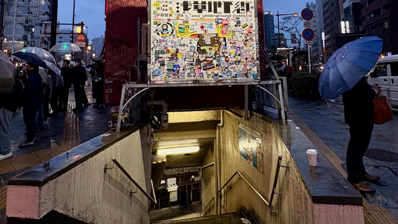 Rainy Tokyo subway entrance with umbrellas and stairs.